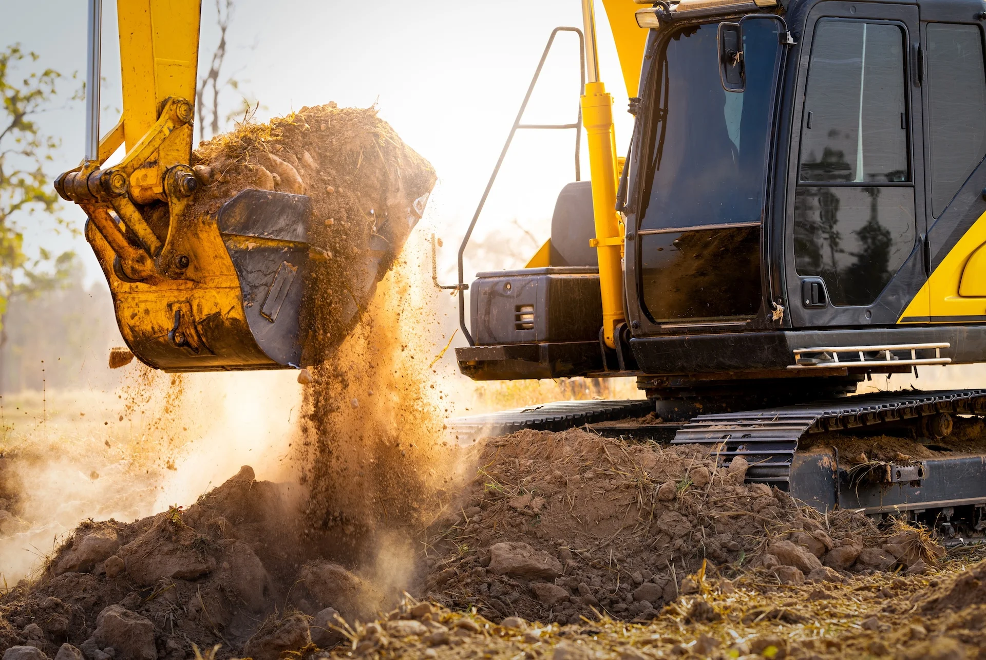 Yellow excavator tipping a bucket of dirt into a spoil pile in golden afternoon light